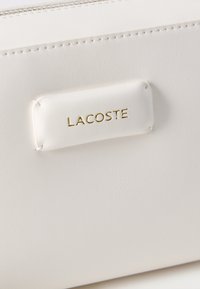 Λευκή επιφάνεια από δέρμα με ραμμένο λογότυπο Lacoste, ανάγλυφο με χρυσά γράμματα.