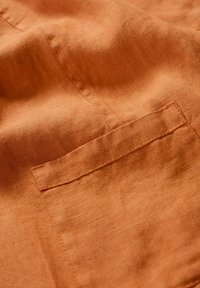 Linen w kolorze rdzy z lekko teksturową powierzchnią, z przeszytą kieszenią po prawej stronie.