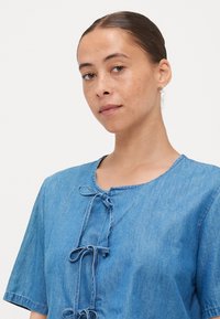 Svetlo modra denim Bluza s kratkimi rokavi, okroglo izrezom in dekorativnimi detajli mašnjice spredaj. Gladka tekstura, sproščen dizajn.