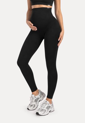 Mujer con leggings negros de cintura alta y zapatillas plateadas New Balance, con una mano apoyada en la cadera y la otra en el estómago.