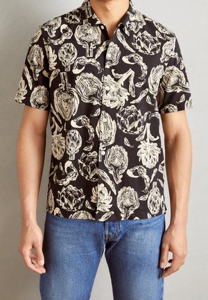 Mann trägt ein schwarzes kurzärmeliges Hemd mit beigefarbenem Artischockenmuster, kombiniert mit blauen Jeans vor neutralem Hintergrund.