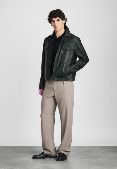 Νεαρός άντρας φορώντας μαύρο δερμάτινο μπουφάν, μαύρο πουκάμισο, μπεζ παντελόνι σε φαρδιά γραμμή, μαύρα παπούτσια και ένα μωβ γάντι στο αριστερό χέρι, που στέκεται μπροστά σε έναν γκρι τοίχο.