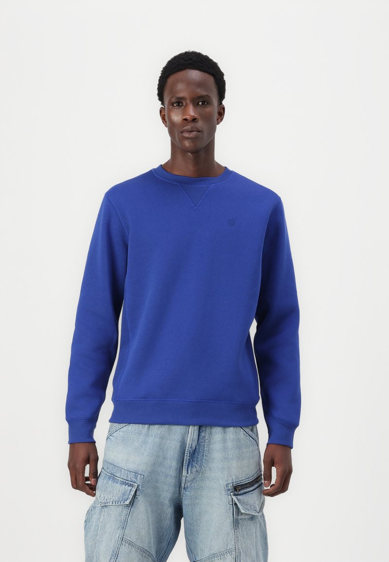 Kék crewneck pulóver puha anyagból, bordázott mandzsettával és szegéllyel, a mellkason egy diszkrét logóval. Világos farmer cargo nadrággal párosítva.