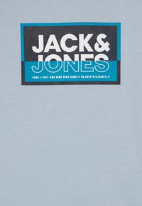Šviesiai mėlyna medvilninė marškinėliai su juodu ir turkio spalvos grafiniu užrašu "JACK & JONES" ir geografinių koordinatų nurodymu.