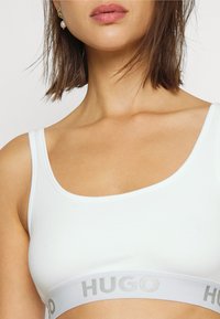 Hvit bomullsbralette med rund hals og brede stropper, prydet med en grå "HUGO" logo-bånd langs bunnen.