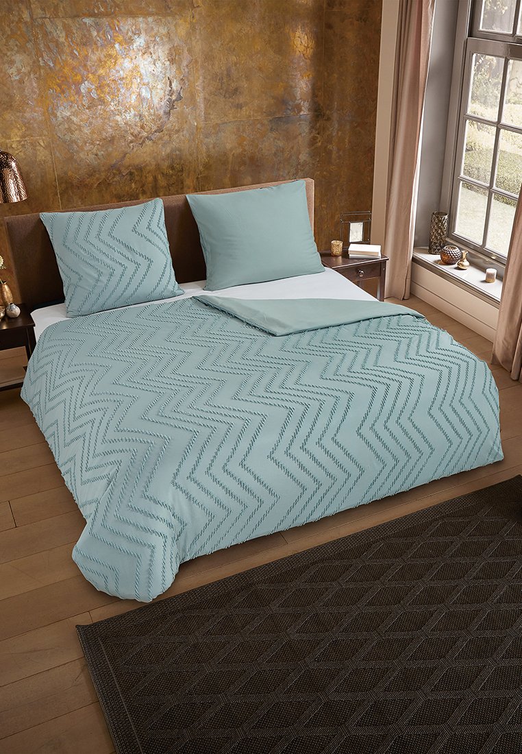 Couverture de lit bleu clair avec un motif en zigzag, accompagnée de taies d'oreiller assorties. Cadre de lit en bois et tapis texturé foncé sur un sol en bois.