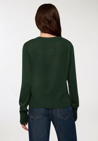 Ciemnozielony sweter dzianinowy z okrągłym dekoltem i prążkowanymi mankietami, o lekko skróconym kroju i teksturowanej tkaninie. W zestawieniu z niebieskimi dżinsami.
