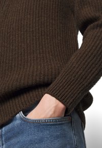 Brązowy sweter z prążkowanego dzianiny z długimi rękawami, w połączeniu z niebieskimi dżinsami. Zbliżenie ukazuje teksturę materiału oraz dłoń w kieszeni.