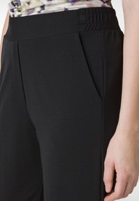 Zwarte broek van gladde, rekbare stof met een brede elastische tailleband en zijzakken. Heeft een strakke, gestroomlijnde uitstraling.