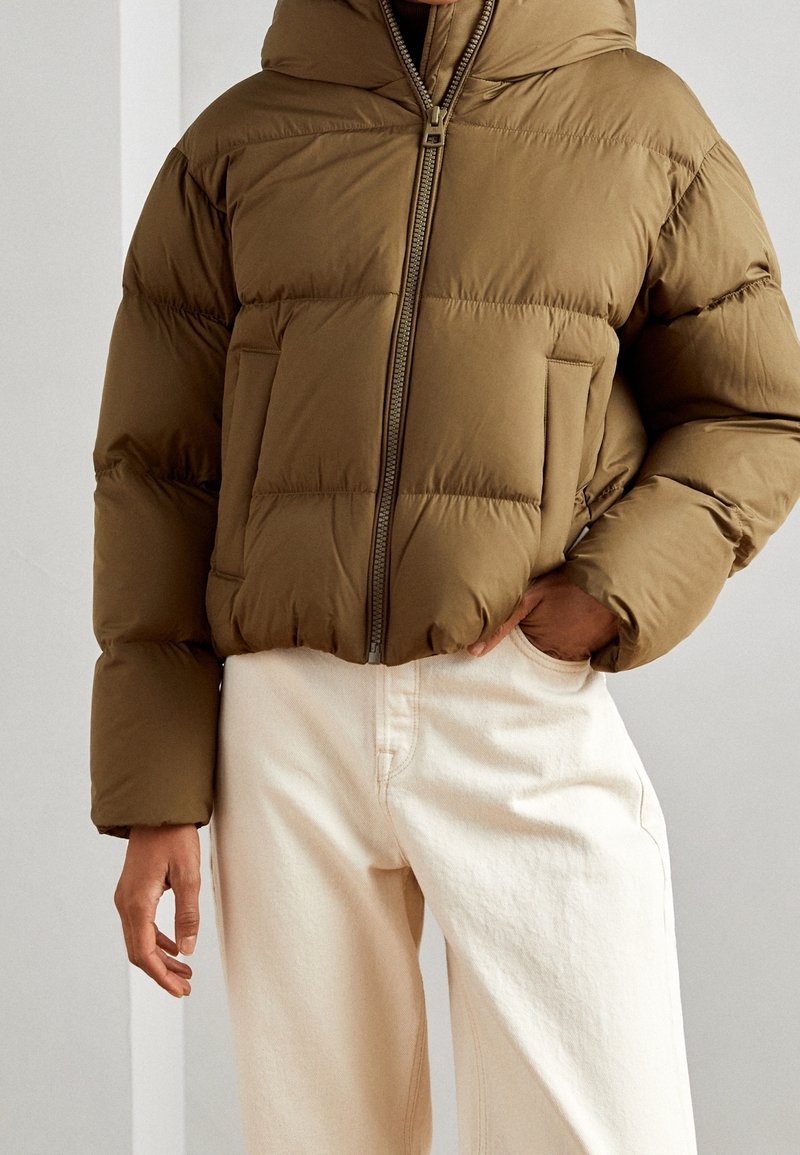Personne portant une doudoune marron zippée avec des poches latérales et un pantalon écru, une main dans la poche et l'autre détendue sur le côté.