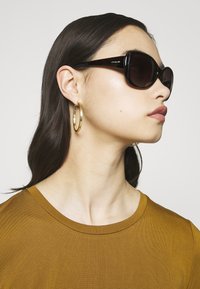 Oversized sunglasses with dark lenses, thick black frames. The model wears gold hoop earrings and a brown crew-neck shirt.