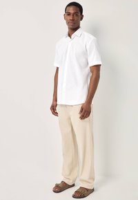 Uomo in piedi che indossa una camicia bianca a maniche corte con bottoni, pantaloni beige larghi e sandali marroni con fibbia su uno sfondo chiaro e uniforme.