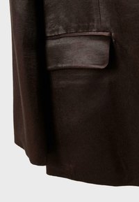 Primo piano di tessuto per indumento marrone scuro che mostra una tasca con patta con una leggera lucentezza e una texture liscia.