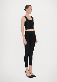Top corto nero con spalline spesse e un accento dorato in vita, abbinato a leggings neri aderenti e tacchi a spillo neri.
