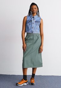 Vest din denim, fustă verde măsliniu cu un brâu elastic și pantofi sport portocalii cu accente negre. Design simplu, textură netedă.