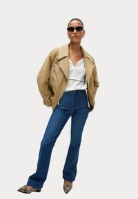 Béžová semišová motorkářská bunda, bílá texturovaná blůza, modré jeansy s vysokým pasem a rozšířenými nohavicemi, a černé lodičky s hadím vzorem a špičatou špičkou. Přehnané sluneční brýle.