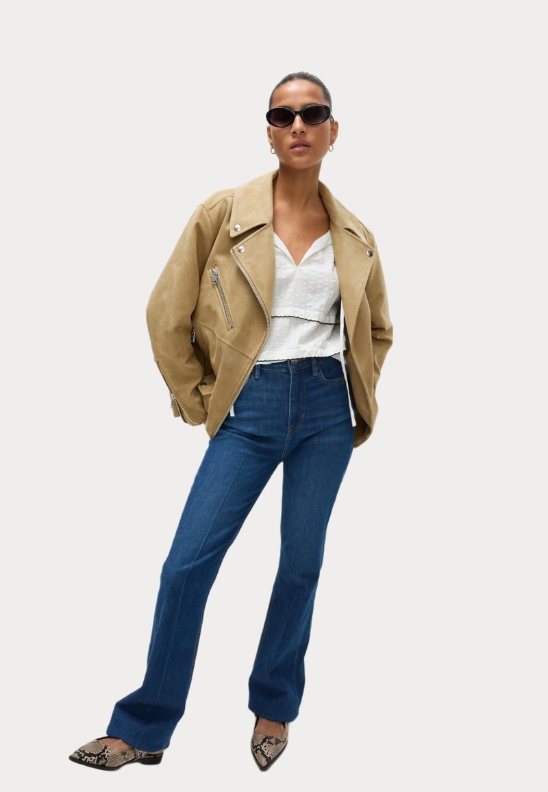 Béžová semišová motorkářská bunda, bílá texturovaná blůza, modré jeansy s vysokým pasem a rozšířenými nohavicemi, a černé lodičky s hadím vzorem a špičatou špičkou. Přehnané sluneční brýle.