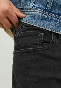 Svarta jeans i denim med subtila sömndetaljer och en framficka, delvis täckta av en ljusblå denimjacka.