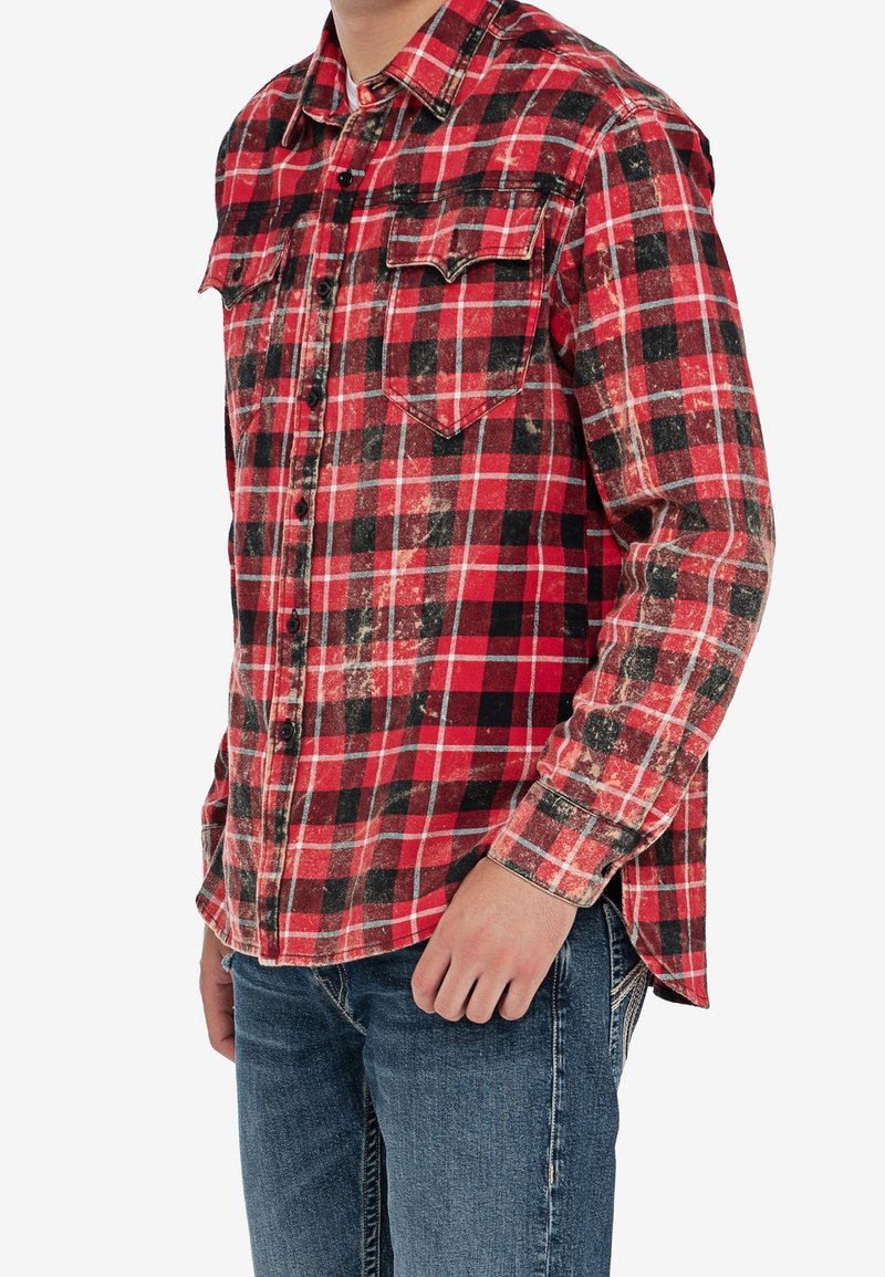 Rood en zwart geruite flanellen shirt met knoopsluiting, twee borstzakken en een licht versleten textuur. Gecombineerd met blauwe jeans.