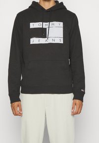 Muž v černé mikině s kapucí s textem "TOMMY JEANS" a obdélníkovým designem, v kombinaci se světle zbarvenými kalhotami, stojící na jednobarevném pozadí.