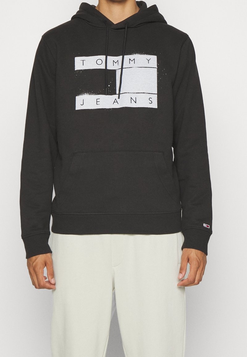 Muž v černé mikině s kapucí s textem "TOMMY JEANS" a obdélníkovým designem, v kombinaci se světle zbarvenými kalhotami, stojící na jednobarevném pozadí.