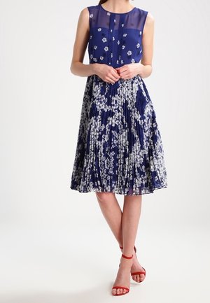 Femme portant une robe bleu marine sans manches à imprimé floral blanc avec une jupe plissée, associée à des escarpins rouges à bride cheville.