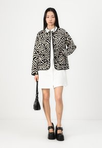 Jachetă cu un model alb și negru, cu un design pătrățos, având două buzunare frontale, purtată peste o rochie albă, asortată cu accesorii negre.
