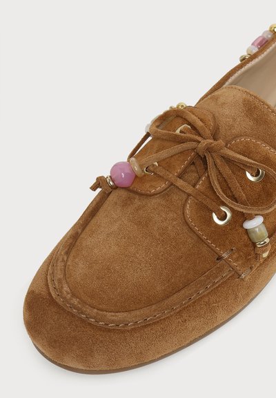 Mocassin en daim marron avec un lacet en forme de nœud papillon et des perles décoratives roses, blanches et dorées près des lacets et de la cheville.