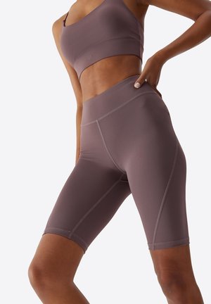 Femme portant un soutien-gorge de sport mauve et un short de vélo taille haute, une main sur la hanche, sur fond clair uni.