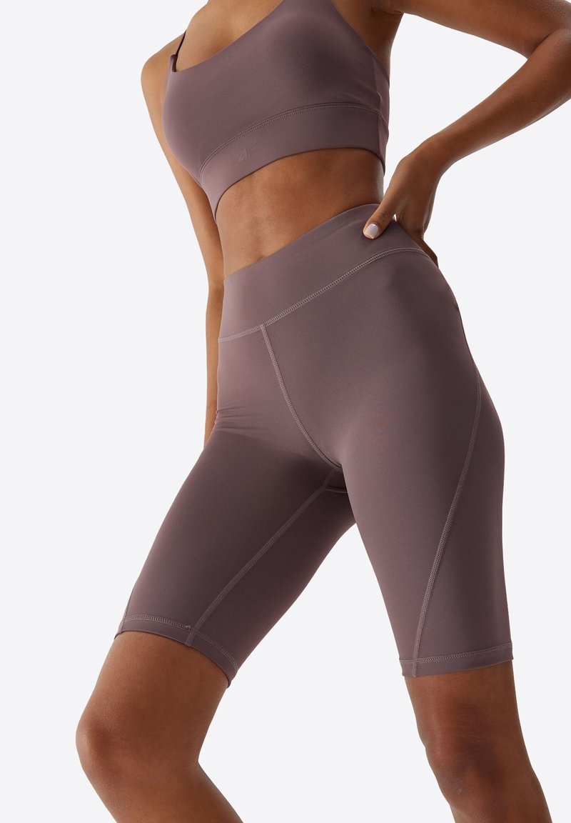 Femme portant un soutien-gorge de sport mauve et un short de vélo taille haute, une main sur la hanche, sur fond clair uni.