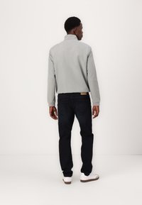 Grå lett jakke med høy krage, kombinert med mørkeblå jeans og hvite sneakers. Teksturen virker glatt; jakken har elastiske mansjetter.