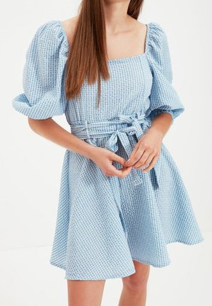 Młoda kobieta ubrana w jasnoniebieską, teksturowaną sukienkę z bufkami i przewiązanym paskiem w talii, stoi z rękami złączonymi z przodu.