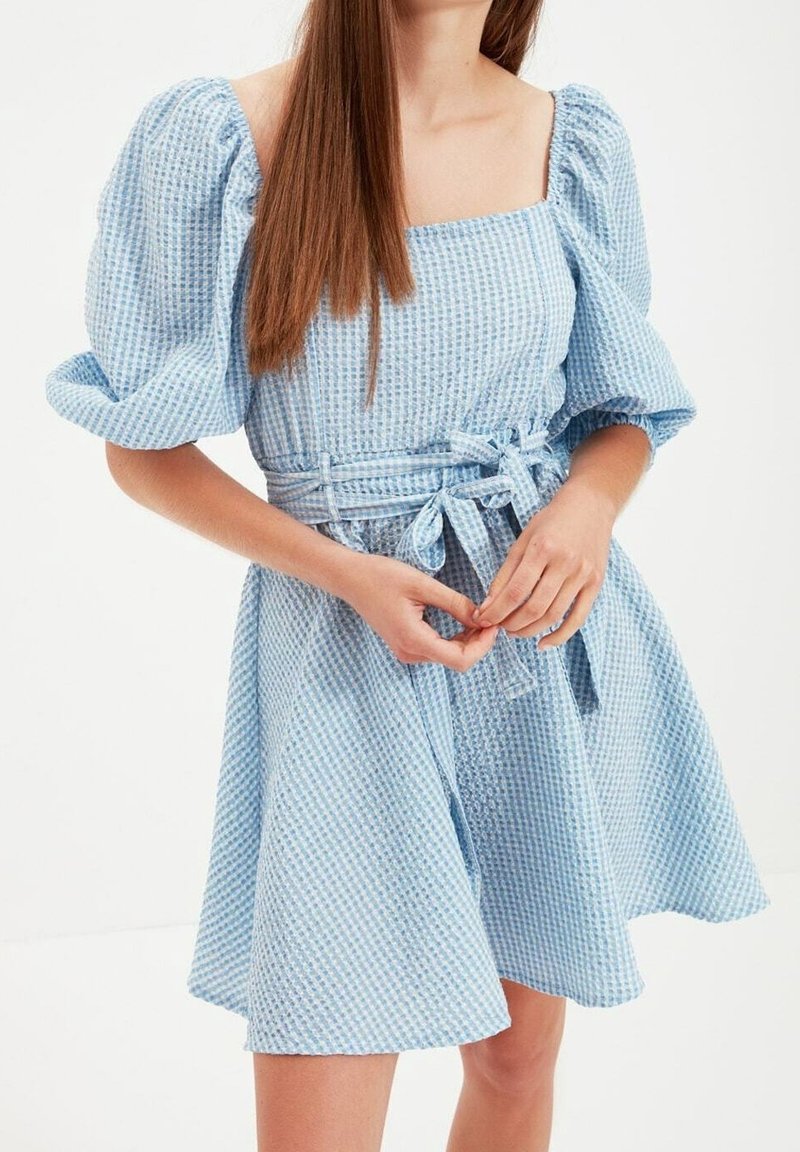 Jeune femme portant une robe bleu clair texturée avec des manches bouffantes et une ceinture nouée à la taille, debout les mains jointes devant elle.
