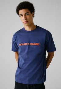 Tamsiai mėlynas medvilninis marškinėlis su oranžine grafika "RED BULL RACING" logotipu ant krūtinės. Trumpomis rankovėmis, apvaliu iškirpimu ir laisvu pritaikymu.