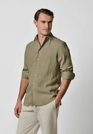 Mand iført en lys olivengrøn skjorte med knapper og opkrogede ærmer samt hvide bukser, stående med den ene hånd i lommen.