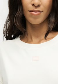 Femme à la peau moyenne foncée et aux cheveux foncés ondulés portant un haut blanc avec un petit logo carré rose clair sur la poitrine.