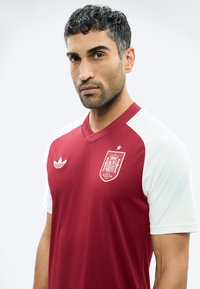 Άνδρας με κοντά σγουρά μαύρα μαλλιά φοράει κόκκινη και λευκή φανέλα της ισπανικής ομάδας ποδοσφαίρου με το λογότυπο της Adidas, κοιτώντας σοβαρά απέναντι σε απλό φόντο.