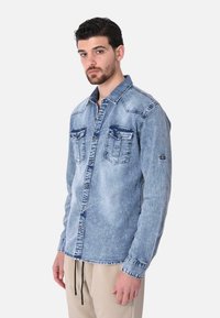 Camicia in denim azzurro chiaro con bottoni, dotata di due tasche sul petto, cuciture a contrasto e vestibilità  rilassata. Maniche lunghe con polsini abbottonati.