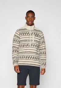 Pull-over en polaire beige avec des motifs géométriques noirs, col zippé mi-hauteur et accent de logo ; associé à un short sombre. Texturé lisse.
