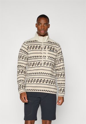 Beige fleece-pulloveilla, joissa on mustia geometrisia kuvioita, puolivälin vetoketjukaulus ja logoväri; yhdistetty tummiin shortseihin. Sileä tekstuuri.