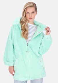 Puha menta zöld cipzáros felső nagy kapucnival, bő szabással és első zsebekkel. Simább, texturált anyagból készült, alatta kontrasztos csíkos ing található.