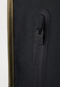 Tissu noir texturé avec une fermeture éclair noire lisse et une languette en tissu. Présente des accents verts le long des bords.