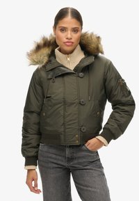 Olijfgroene, cropped bomberjack met een nepbont capuchon, knoopsluitingen en geribbelde boorden, gedragen over een lichtbeige gebreide trui.