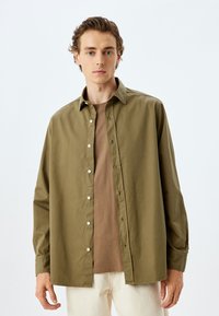 Oliivinvihreänapitettava paita rentoa istuvuutta, pitkät hihat ja valkoiset napit, päälle puettuna beige crew-neck t-paita.