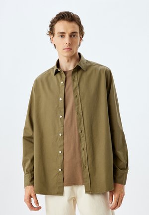 Olivengrøn knappet skjorte med løs pasform, lange ærmer og hvide knapper, lagd over en beige crew-neck T-shirt.