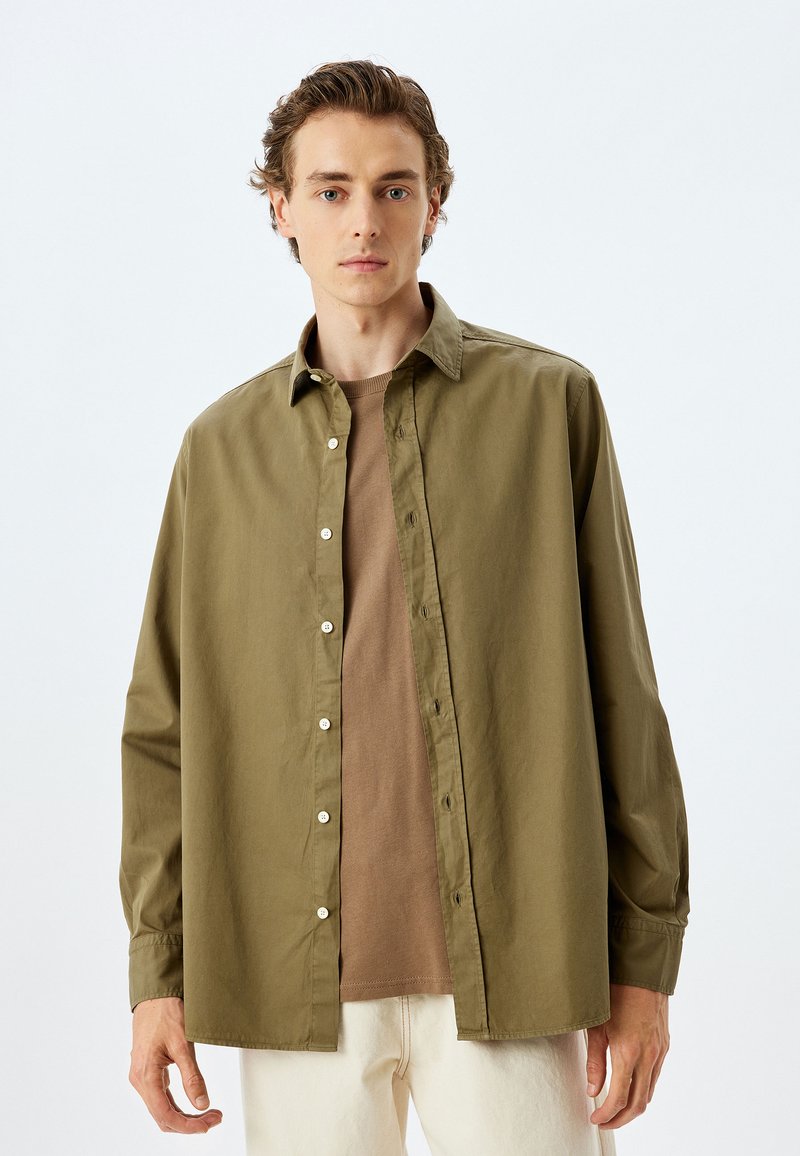 Oliivinvihreänapitettava paita rentoa istuvuutta, pitkät hihat ja valkoiset napit, päälle puettuna beige crew-neck t-paita.