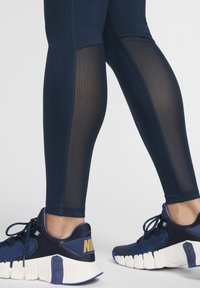 Leggings sportivi blu navy con pannelli in rete e cuciture a contrasto, abbinati a scarpe da ginnastica blu e bianche con un logo prominente e suola testurizzata.