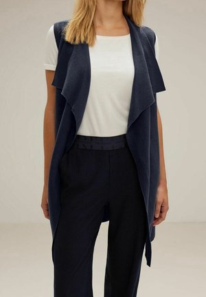 Mujer con una camiseta blanca de manga corta, cárdigan sin mangas azul marino drapeado y pantalones negros de tiro alto, de pie frente a un fondo liso.