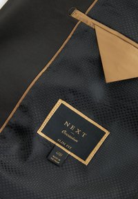 Tissu noir texturé avec bordure marron et une étiquette indiquant "NEXT Occasion Slim Fit 40R Made in Vietnam."