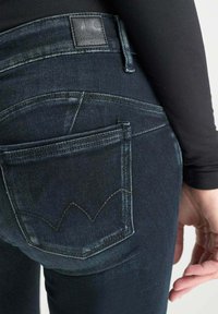 Jeans de mezclilla oscura con una textura suave. Presentan un diseño distintivo en el bolsillo trasero y una etiqueta de cuero en la cinturilla.